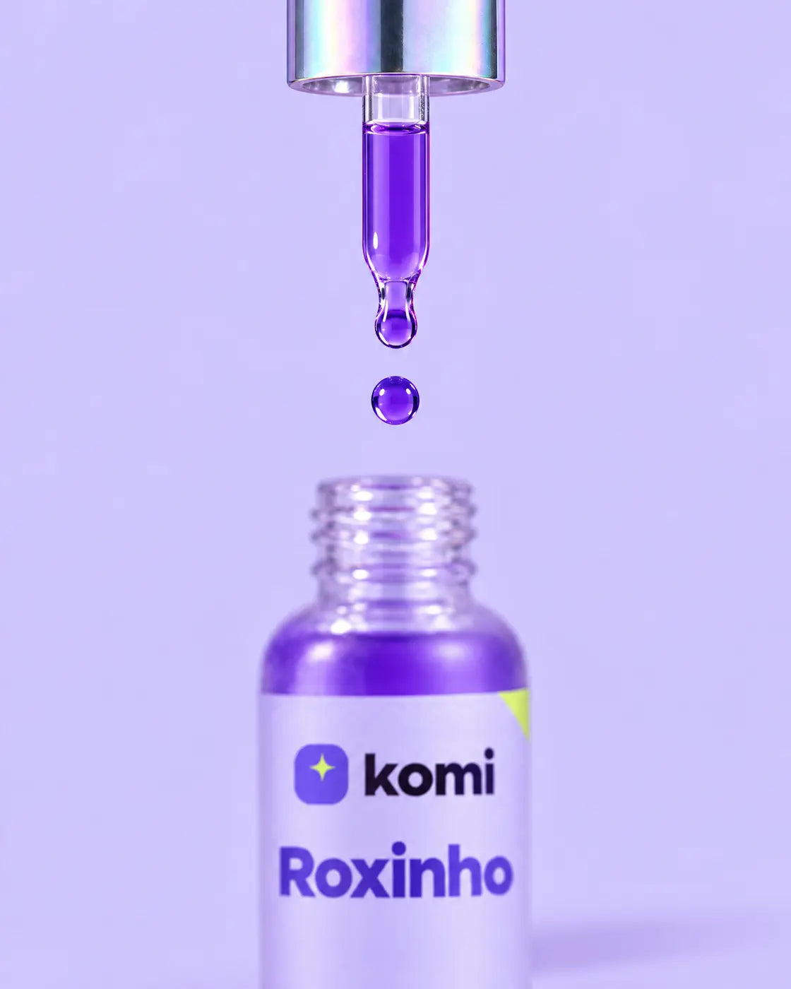 Gota roxa caindo da pipeta cromada sobre o frasco aberto do Roxinho, corretor de cor Uva Roxa de 30 ml, em fundo lavanda
