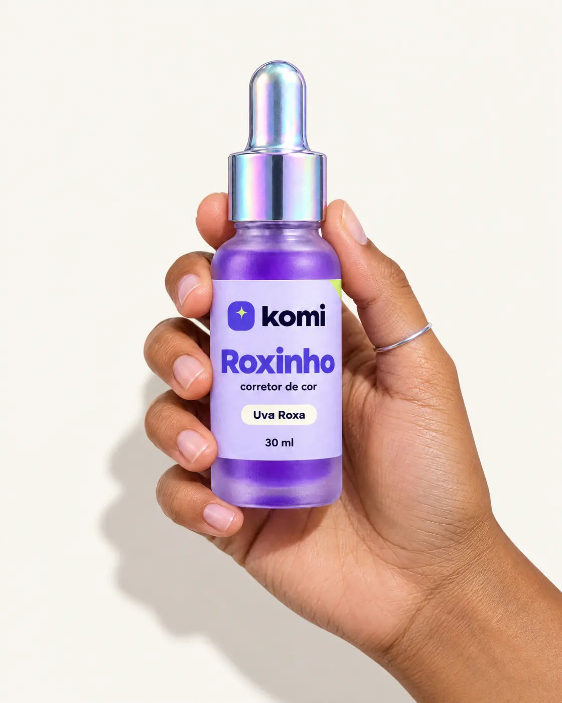 Mão segurando o frasco do Roxinho, corretor de cor Uva Roxa de 30 ml, em fundo leite